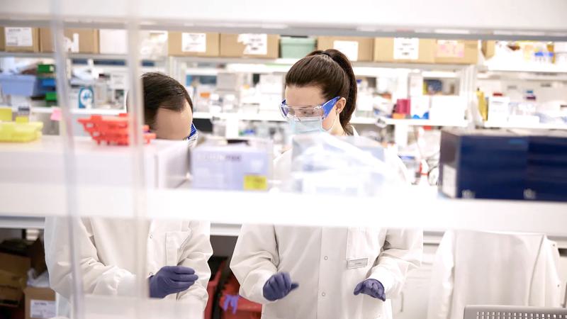 Two scientists in lab coats and goggles stand at a lab bench talking with each other. A shelf with lab materials is out of focus in the foreground.