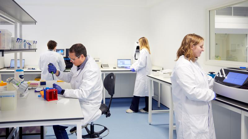 Four scientists in lab coats working in a lab space. One scientist sits at a lab bench micropipetting samples. One scientist stands next to a machine. The two other scientists stand in the background working at computers.