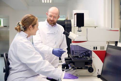 Two scientists in lab coats working together in the lab while one sits at a microscope and the other stands next to the machine.